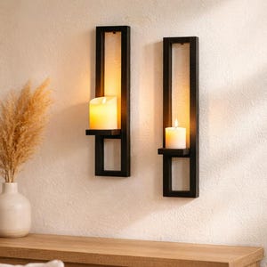 Può includere: Due applique nere con candele accese. Ogni applique ha un design rettangolare con un ripiano per tenere la candela. Un vaso con erba ornamentale secca è posto su un tavolo di legno.