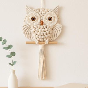 Op de afbeelding: Een wandhanger in de vorm van een crèmekleurige macramé uil met grote bruine ogen, zittend op een horizontale houten stok. De uil is gemaakt van geknoopt koord met een gestructureerd borstpatroon en een lange, rechte franje aan de onderzijde.