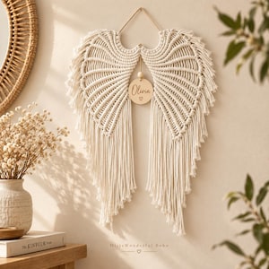 Op de afbeelding: Een crèmekleurige macramé wandhanger in de vorm van engelvleugels. Het stuk is voorzien van ingewikkelde geknoopte koordpatronen en lange franjes, met een rond houten label in het midden gegraveerd met de naam Olivia en een hartje.