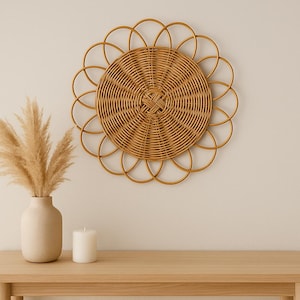 Può includere: Una decorazione da parete decorativa in rattan intrecciato con un design a forma di sole. Un vaso beige con erba della pampa essiccata e una candela bianca sono su un tavolo di legno chiaro. Il muro è dipinto di un beige neutro.