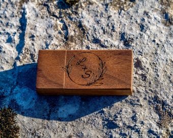 Op de afbeelding: Een rechthoekige houten doos met een donkerbruine afwerking. De doos heeft een lasergeëtst ontwerp van een cirkelvormige krans met de letters "L + S". De doos ligt op een getextureerd stenen oppervlak.