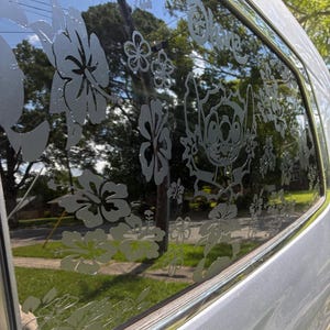 以下が含まれることがあります： 銀色の車の窓に、ハイビスカスの花、その他の花の模様、キャラクターのスティッチがエッチングされています。「Ohana」という言葉もガラスにエッチングされています。背景には木と青い空が写っています。