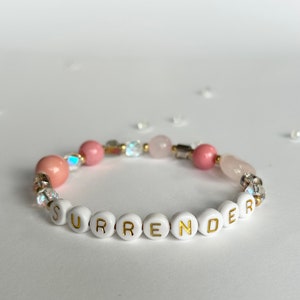 May include: A pink and white beaded bracelet with the word "SURRENDER" spelled out in gold letters on white beads.