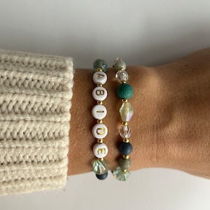 May include: Two bracelets with green, blue, and clear beads. One bracelet has white beads with the word "ABIDE" spelled out in gold letters.