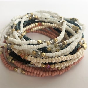 May include: A stack of beaded bracelets in shades of white, pink, black, and gold. The bracelets are made with small, round beads and feature gold and crystal accents.