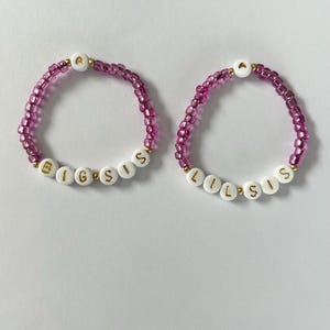 May include: Two beaded bracelets with the words "BIG SIS" and "LIL SIS" spelled out in white beads with gold lettering. The bracelets are pink and have a gold heart charm.