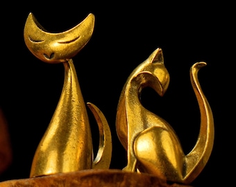 Può includere: Due statuette di gatto color oro. Un gatto ha un corpo alto e snello con una testa a forma di mezzaluna. L'altro gatto è seduto con la coda curva. Entrambi hanno una finitura metallica liscia e sono esposti su una superficie marrone.