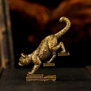 May include: A detailed, golden-colored cat figurine climbing a set of steps. The cat is in mid-motion, with its tail curved upwards. The steps are rectangular and appear to be made of the same material as the cat. The background is dark and blurred.