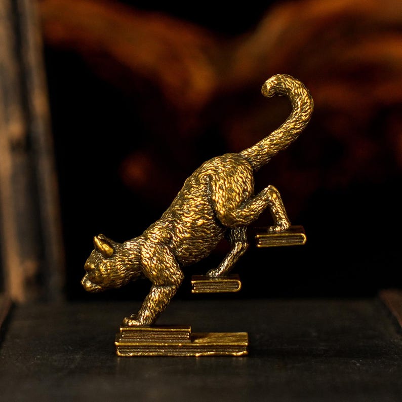 May include: A detailed, golden-colored cat figurine climbing a set of steps. The cat is in mid-motion, with its tail curved upwards. The steps are rectangular and appear to be made of the same material as the cat. The background is dark and blurred.