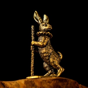 May include: A golden-colored rabbit figurine with a ruffled collar and a staff. The rabbit is standing upright on a wooden surface against a black background. The figurine has detailed fur and a regal appearance.