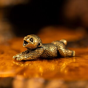 May include: A small, metallic, gold-toned figurine of a sloth in a prone, stretching position. The sloth has a textured surface mimicking fur, with its front paws extended forward and its mouth open in a wide, yawning expression.