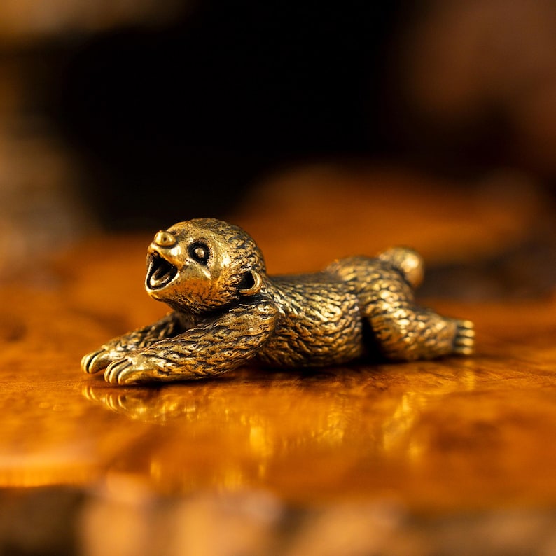 May include: A small, metallic, gold-toned figurine of a sloth in a prone, stretching position. The sloth has a textured surface mimicking fur, with its front paws extended forward and its mouth open in a wide, yawning expression.