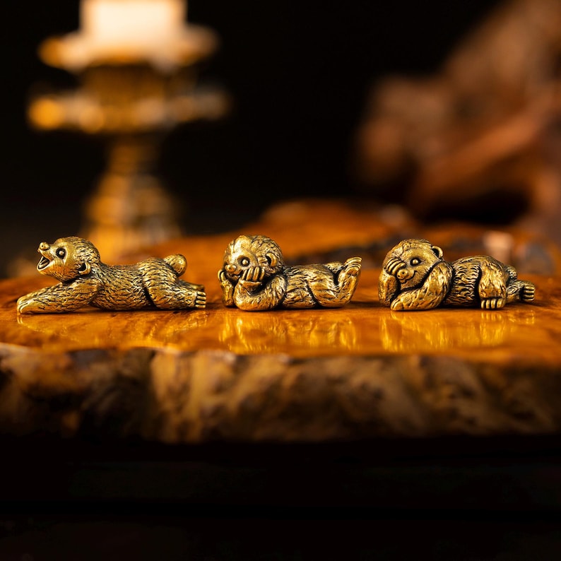 May include: Three small, metallic gold-toned animal figurines in a reclining position. The first figure is lying flat with its head raised, the second rests its head on one hand, and the third covers its eyes with its paws, all featuring textured fur.