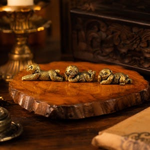 May include: Three small, metallic, gold-toned monkey figurines arranged in a row. Each monkey is depicted in a different pose: one reaching forward, one resting its head on its hand, and one covering its eyes with its hands, all featuring textured fur details.