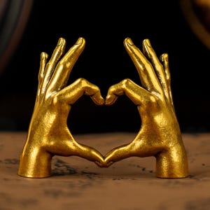 Puede incluir: Una escultura dorada de dos manos formando un corazón. Las manos están detalladas con nudillos y líneas visibles, y la escultura tiene un brillo metálico. El fondo es oscuro, enfatizando el color dorado.