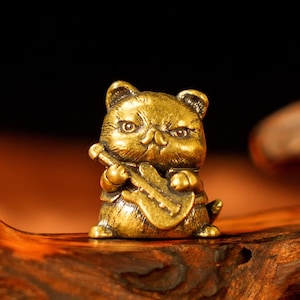 May include: A small, detailed brass figurine of a cat playing a guitar. The cat has an expressive face and is holding the guitar with both paws. The figurine is golden in color and sits on a wooden surface. The background is dark.