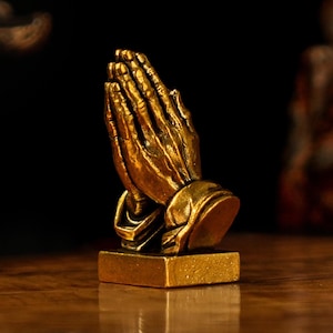 May include: A gold-coloured sculpture of hands clasped in prayer. The hands are detailed with visible knuckles and lines, resting on a rectangular base. The sculpture is set against a dark background.