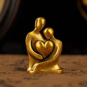 Puede incluir: Pequeña escultura dorada que representa dos figuras abrazándose, con un corazón en el centro. Las figuras son estilizadas y lisas, con un acabado metálico cálido. La escultura es un símbolo de amor y conexión.