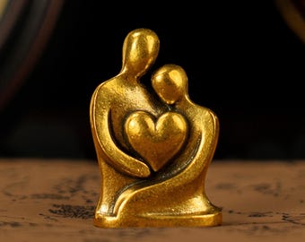 Puede incluir: Pequeña escultura dorada que representa dos figuras abrazándose, con un corazón en el centro. Las figuras son estilizadas y lisas, con un acabado metálico cálido. La escultura es un símbolo de amor y conexión.