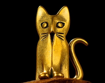 May include: A golden cat figurine with a sitting pose, featuring a curved tail and detailed facial features. The cat is perched on a brown, textured surface against a black background. The figurine's design includes whiskers and pointed ears.