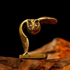 May include: A small, detailed brass owl figurine in flight. The owl has outstretched wings and a focused gaze, with intricate feather detailing. The figurine is mounted on a rectangular base, showcasing a vintage aesthetic.