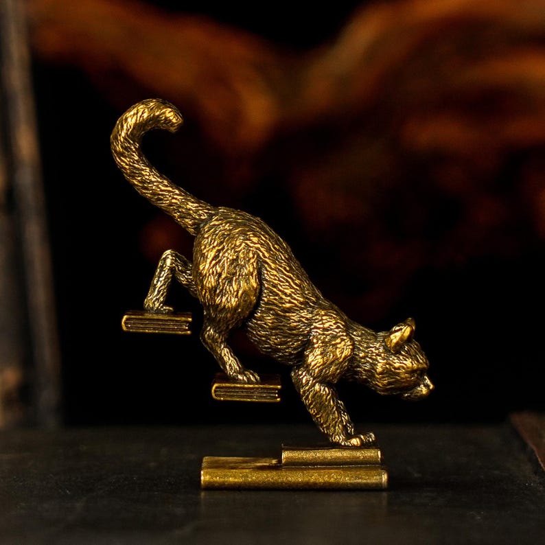 May include: A detailed brass cat figurine climbing a set of miniature steps. The cat is posed mid-climb, with its tail arched. The steps are also made of brass, and the background is a blurred dark brown.