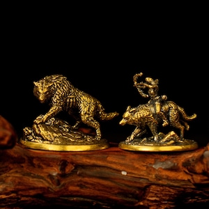 May include: Two small, detailed brass figurines. One depicts a wolf, the other a figure riding a wolf and holding a bow. The figurines are on a wooden surface with a dark background. The wolf figurine is on a rock.