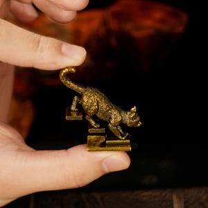 May include: A small, detailed brass cat figurine climbing a set of stairs. The cat is golden-colored with textured fur, and the stairs are rectangular. The figurine is held in a person's hand against a dark background.
