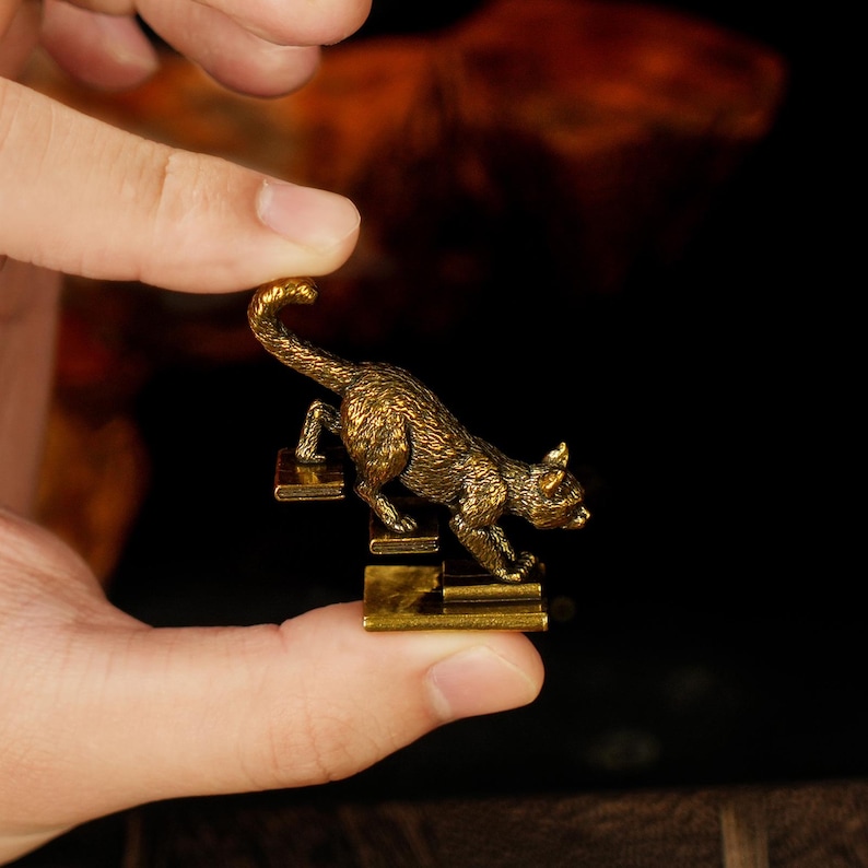 May include: A small, detailed brass cat figurine climbing a set of stairs. The cat is golden-colored with textured fur, and the stairs are rectangular. The figurine is held in a person's hand against a dark background.