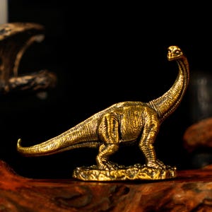 May include: A detailed, golden-colored Brachiosaurus figurine. The dinosaur has a long neck and tail, with textured skin and is standing on a small base. The figurine is set against a dark background, highlighting its metallic finish.