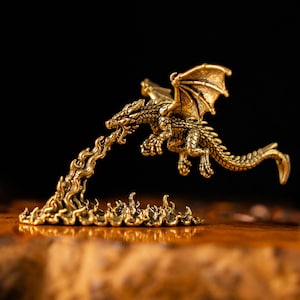 May include: A small, metallic gold-colored dragon figurine in flight, featuring detailed scales and wings. The dragon is depicted exhaling a stream of fire that flows downward to form a textured, flame-shaped base, creating a continuous sculpture.