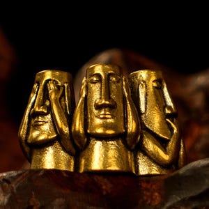 Könnte beinhalten: Drei goldfarbene Statuen in einer Reihe, die jeweils ein stilisiertes Gesicht mit Händen zeigen, die Augen, Ohren und Mund bedecken. Die Statuen stehen auf einer dunklen Holzoberfläche mit dunklem Hintergrund.
