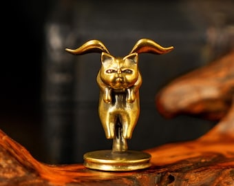 May include: A small, golden cat figurine with large, curved ears and a grumpy expression. The cat is standing upright, holding its paws up to its face. The figurine is mounted on a round base and is made of a shiny, metallic material.