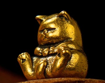 May include: A small, golden cat figurine in a seated position. The cat has a rounded body, small paws, and a slightly grumpy expression. The figurine is made of a shiny, metallic material and sits on a wooden surface against a black background.