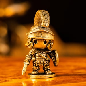 May include: A small, metallic gold-toned figurine of a Roman soldier wearing a crested helmet and segmented armor. The figure holds a short sword in the right hand and a round shield in the left hand, standing on a circular base with a weathered finish.