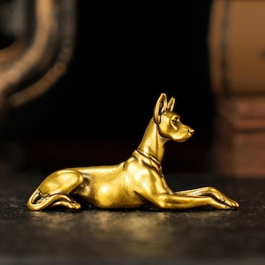 May include: A golden-colored, reclining dog figurine. The dog is lying down with its front paws extended, ears perked, and a collar around its neck. The sculpture has a smooth, polished finish and is set against a dark background.