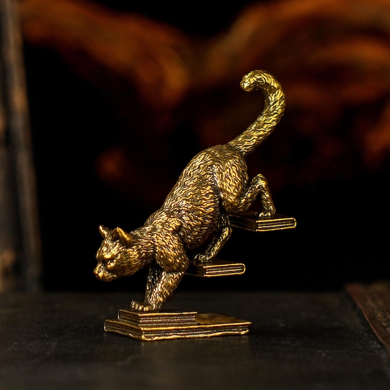 May include: A detailed brass cat figurine in a climbing pose. The cat is depicted with textured fur and is positioned as if scaling a set of steps. The sculpture has a golden hue and is set against a dark background.