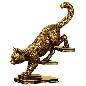 May include: A bronze-colored cat figurine climbing steps made of books. The cat is in a dynamic pose, with its tail arched and body angled upwards. The figurine has a textured surface and is set against a white background.