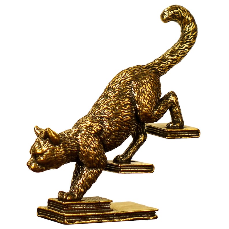 May include: A bronze-colored cat figurine climbing steps made of books. The cat is in a dynamic pose, with its tail arched and body angled upwards. The figurine has a textured surface and is set against a white background.