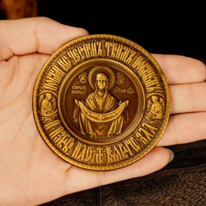 May include: A bronze-toned, circular religious icon held in a hand. The icon features a central figure with a halo, surrounded by text and two angel figures. The design is detailed with intricate patterns and lettering, suggesting a historical or spiritual significance.