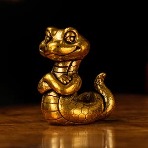 Könnte beinhalten: Goldene Figur einer lächelnden Schlange mit verschränkten Armen. Die Schlange hat einen detaillierten, strukturierten Körper. Die Figur ist wahrscheinlich aus Metall und steht auf einer Holzoberfläche.