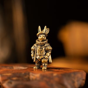 May include: A small brass figurine of an anthropomorphic rabbit dressed in historical attire, including a feathered cap, a ruffled collar, and a tunic with a crescent moon emblem. The rabbit stands upright with detailed clothing textures.