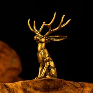 Op de afbeelding: Een messing sculptuur in goudkleur van een jackalope, een mythisch wezen met het lichaam van een haas en het gewei van een hert. De jackalope zit rechtop op een houten voet, met opstaande oren en geweien omhoog. De sculptuur heeft een gouden afwerking.