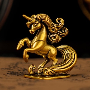 Könnte beinhalten: Goldene Einhornfigur in aufbäumender Pose. Die Skulptur hat eine detaillierte Mähne und einen Schweif sowie ein markantes Horn. Die Figur besteht aus einem glänzenden, metallischen Material und steht auf einem rechteckigen Sockel. Der Hintergrund ist dunkel und hebt die goldene Farbe hervor.
