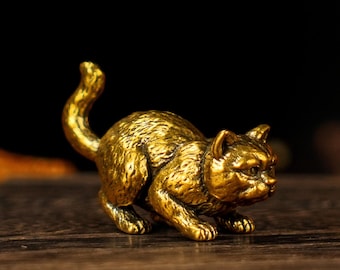 May include: A small, golden cat figurine in a crouching position, with its tail curved upwards. The cat has detailed features and is made of a shiny, metallic material. The figurine is displayed on a wooden surface.