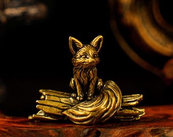 May include: A small, detailed brass fox figurine sitting atop a stylized, textured base. The fox has pointed ears and a bushy tail, with intricate detailing on its fur. The figurine is a warm, golden color.
