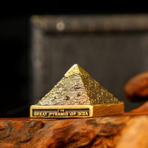 Op de afbeelding: Een gedetailleerde, goudkleurige miniatuur van de Grote Piramide van Gizeh. De piramide heeft een getextureerd oppervlak en een rechthoekige basis met de tekst "THE GREAT PYRAMID OF GIZA". Het is waarschijnlijk een verzamelobject of souvenir.