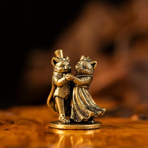 May include: A small brass-colored metal figurine depicting two cats dancing together. One cat wears a top hat and a formal tailcoat, while the other cat wears a crown and a long, textured gown. The figures are joined at the paws on a circular base.