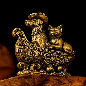 Op de afbeelding: Een goudkleurige metalen sculptuur van een hond met een hoed en een kat in een sierlijke boot. De boot heeft ingewikkeld krulwerk en sterdetails tegen een donkere achtergrond.