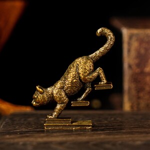 May include: A detailed, golden-colored cat figurine climbing a set of miniature steps. The cat is in a dynamic pose, with its tail curved upwards. The figurine is made of metal and has a textured surface, suggesting a bronze or brass material.